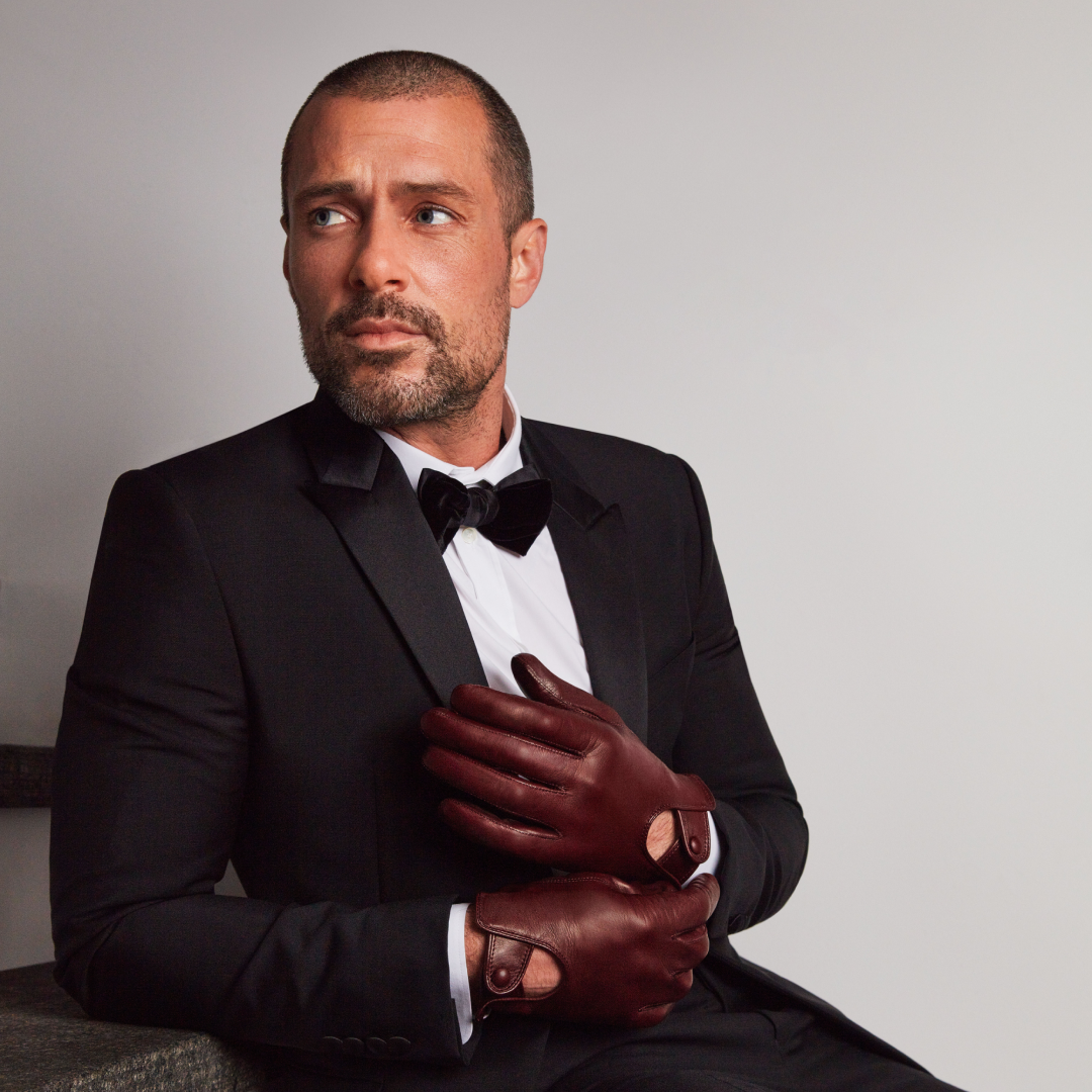 A well-dressed man in a black tuxedo and bow tie wearing burgundy leather driving gloves with a snap-button closure, exuding elegance and sophistication.