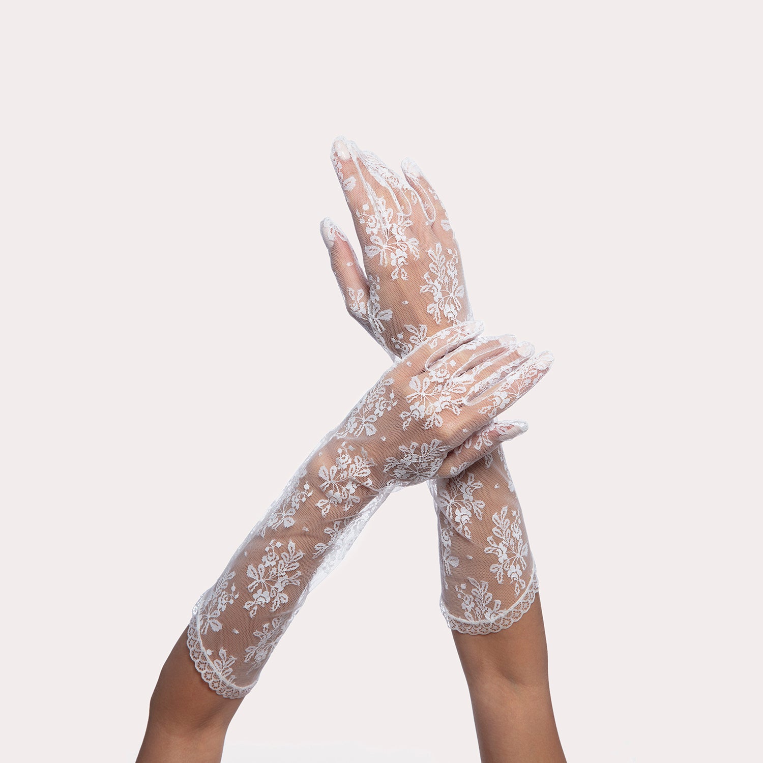 Close-up of crossed hands wearing sheer white floral lace gloves, elegantly posed against a light background, showcasing delicate bridal accessory details.