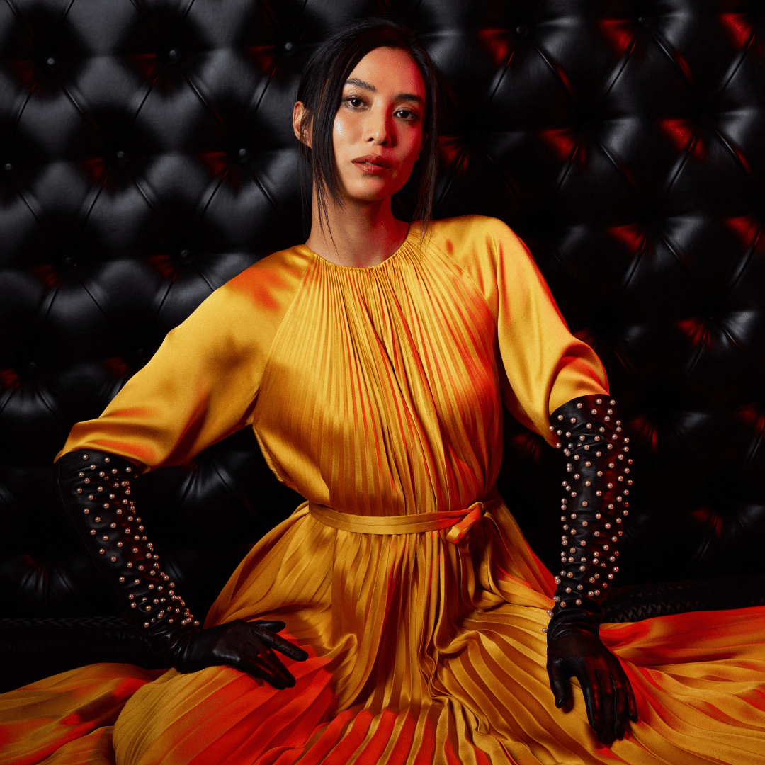 Stylish woman in a golden pleated satin gown and black opera gloves embellished with large pearls, seated against a black tufted leather backdrop.