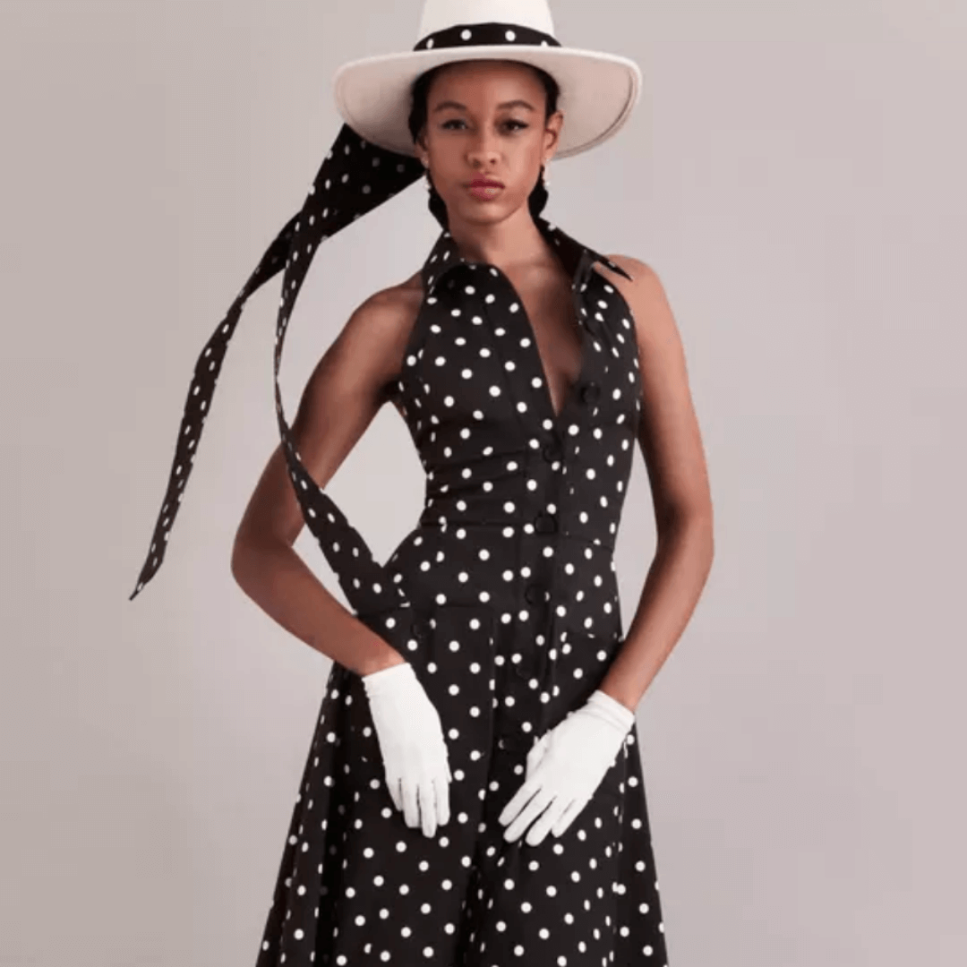 Stylish woman in a black and white polka dot halter dress, white gloves, and a wide-brim hat with trailing ribbons, posing against a neutral backdrop.
