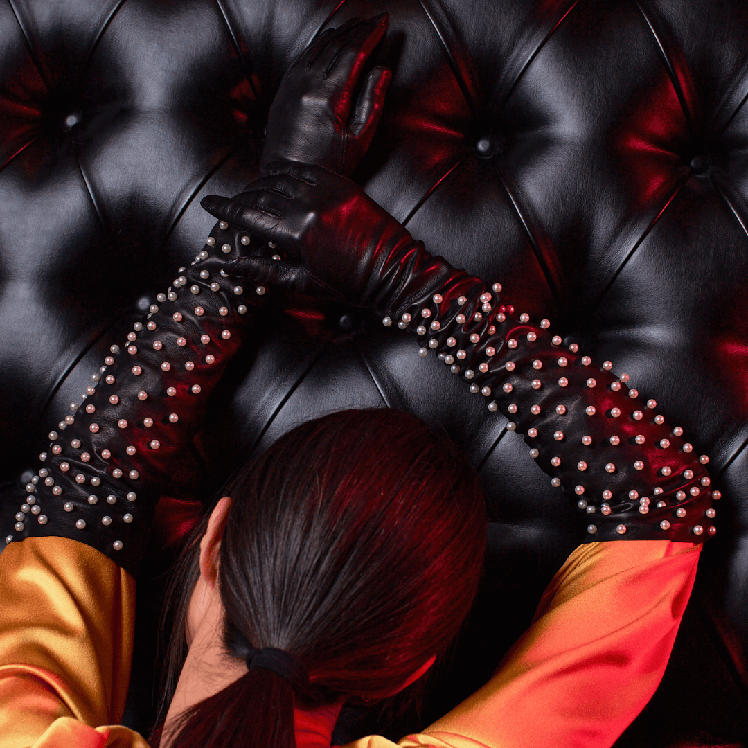 Back view of a woman with a sleek ponytail wearing black leather opera gloves adorned with large pearls, leaning against a tufted black leather wall.