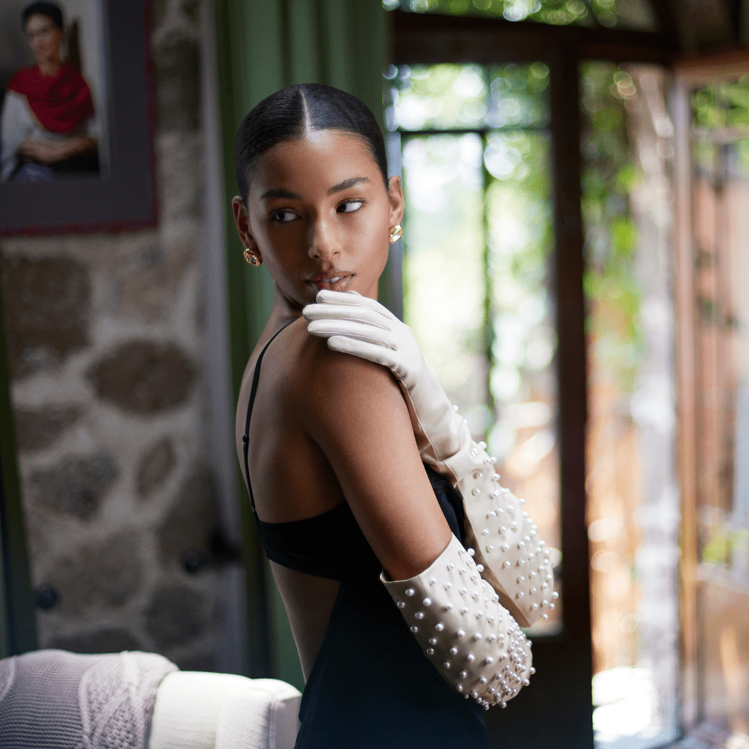 Elegant woman wearing bone color opera gloves adorned with large pearls, styled with a black evening dress and gold earrings in a softly lit, refined interior.