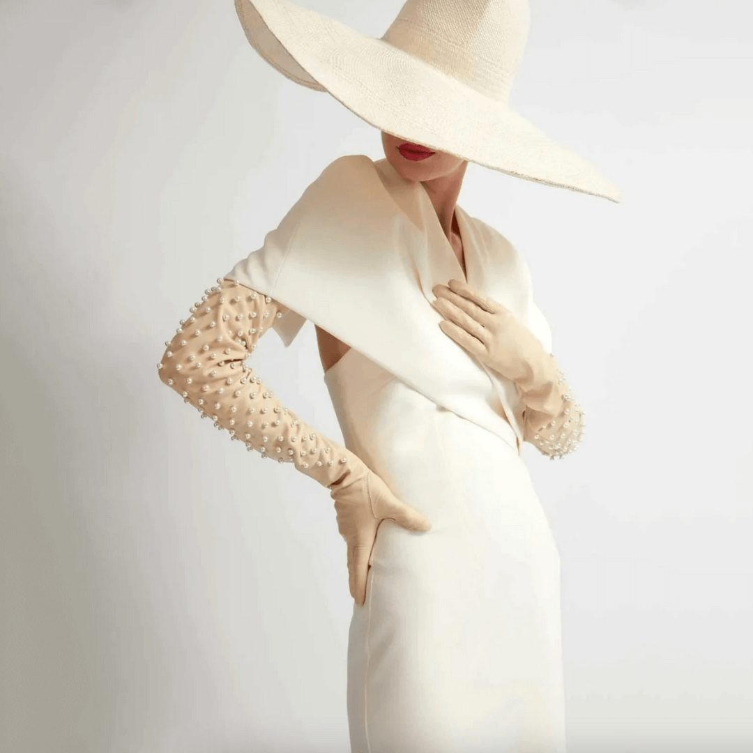 Fashion model in an ivory gown and oversized sun hat wearing bone colored leather opera gloves embellished with pearls, striking a poised, minimalist pose.