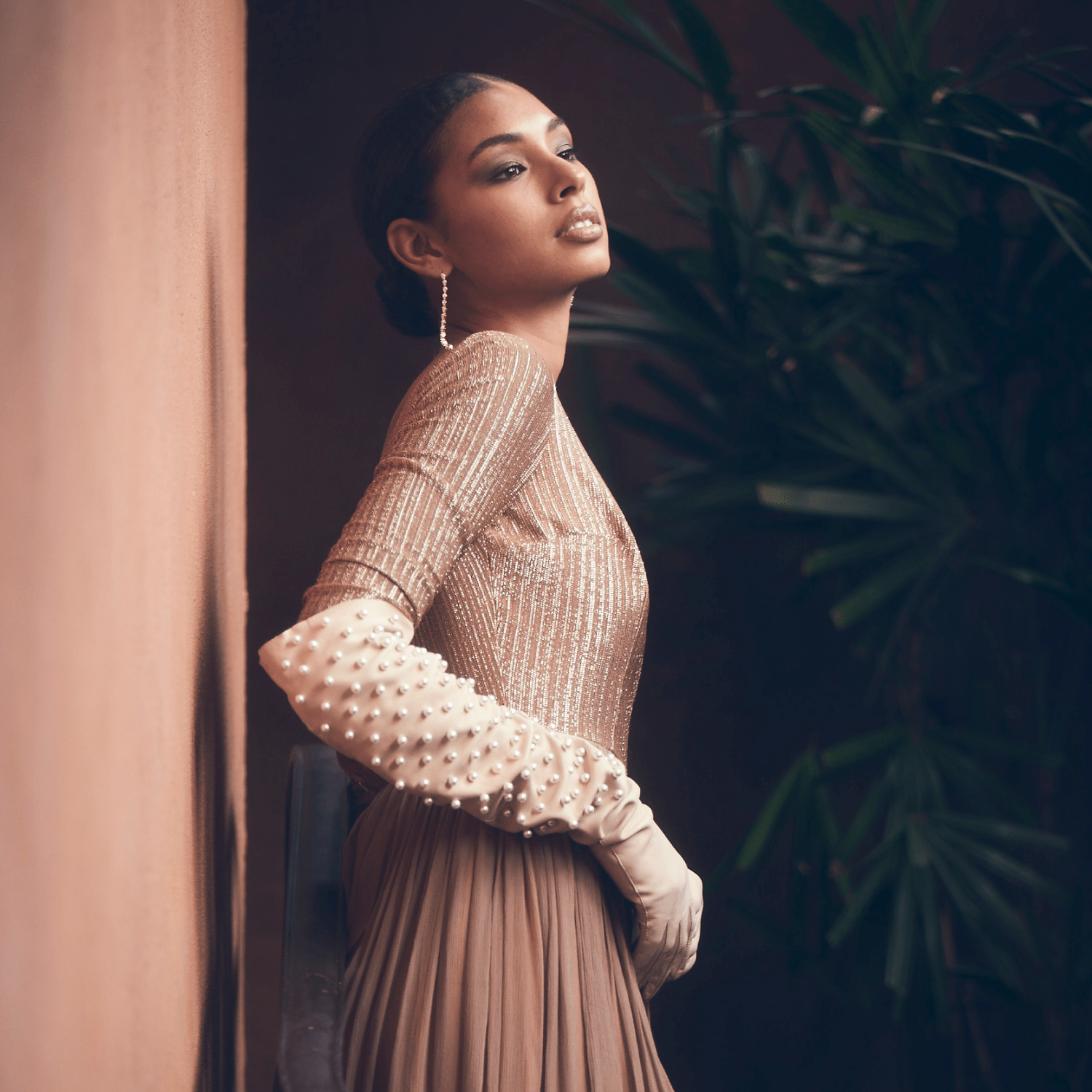 Elegant woman wearing pearl-embellished bone colored opera gloves and a gold sequin top with pleated skirt, posing gracefully against a warm-toned wall and greenery.