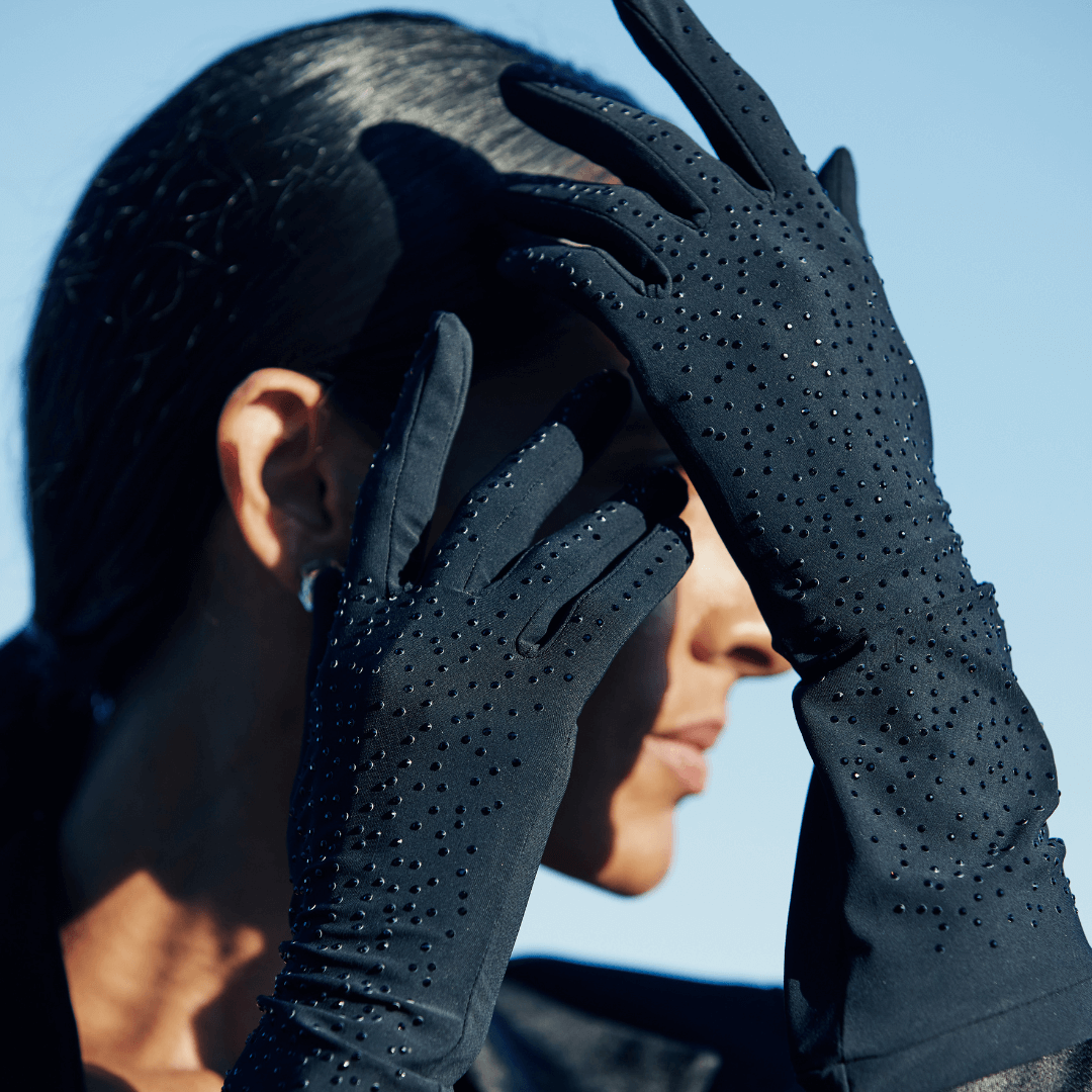 Close-up of a woman wearing black crystal-embellished gloves, holding her hands over her face against a clear blue sky background.