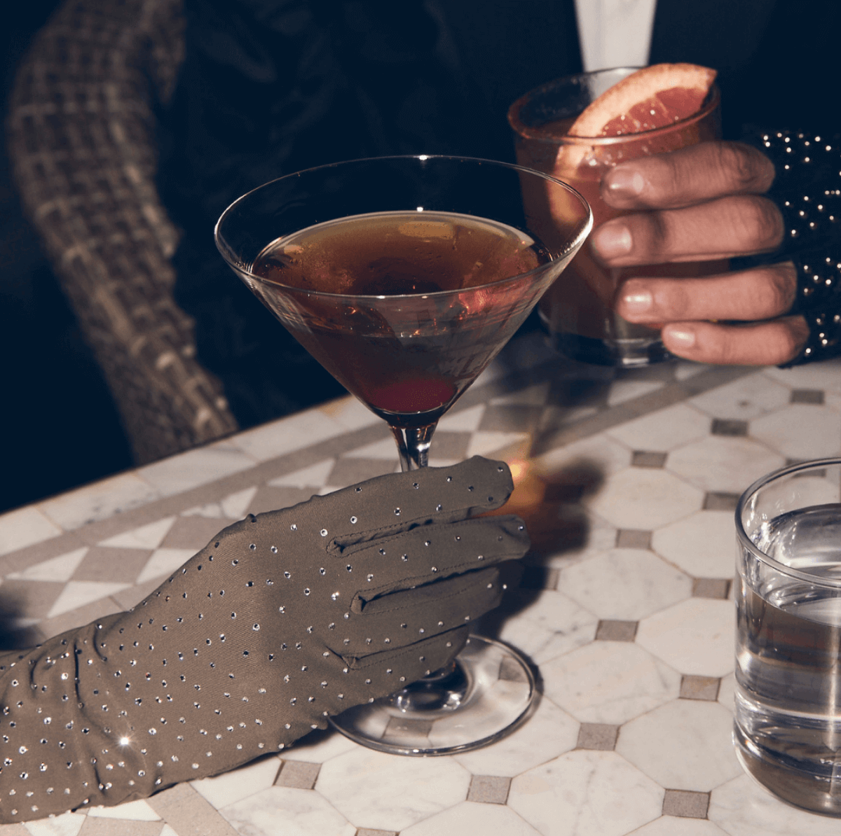 Gloved hands holding cocktails at a marble table—one wearing crystal-studded gloves holding a martini, the other with a whiskey glass garnished with grapefruit.