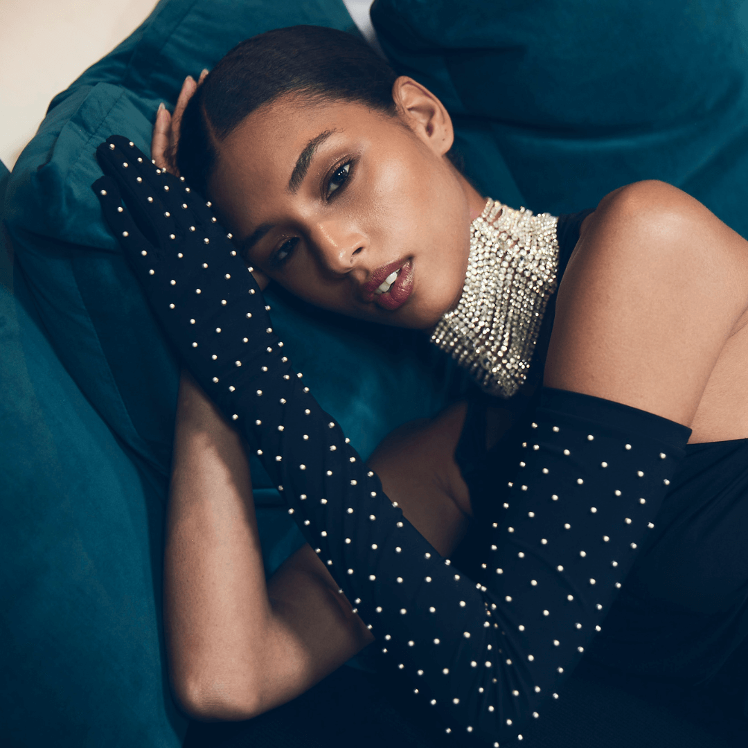 Elegant woman reclining on a velvet sofa wearing long black opera gloves adorned with pearls and a statement pearl choker necklace.