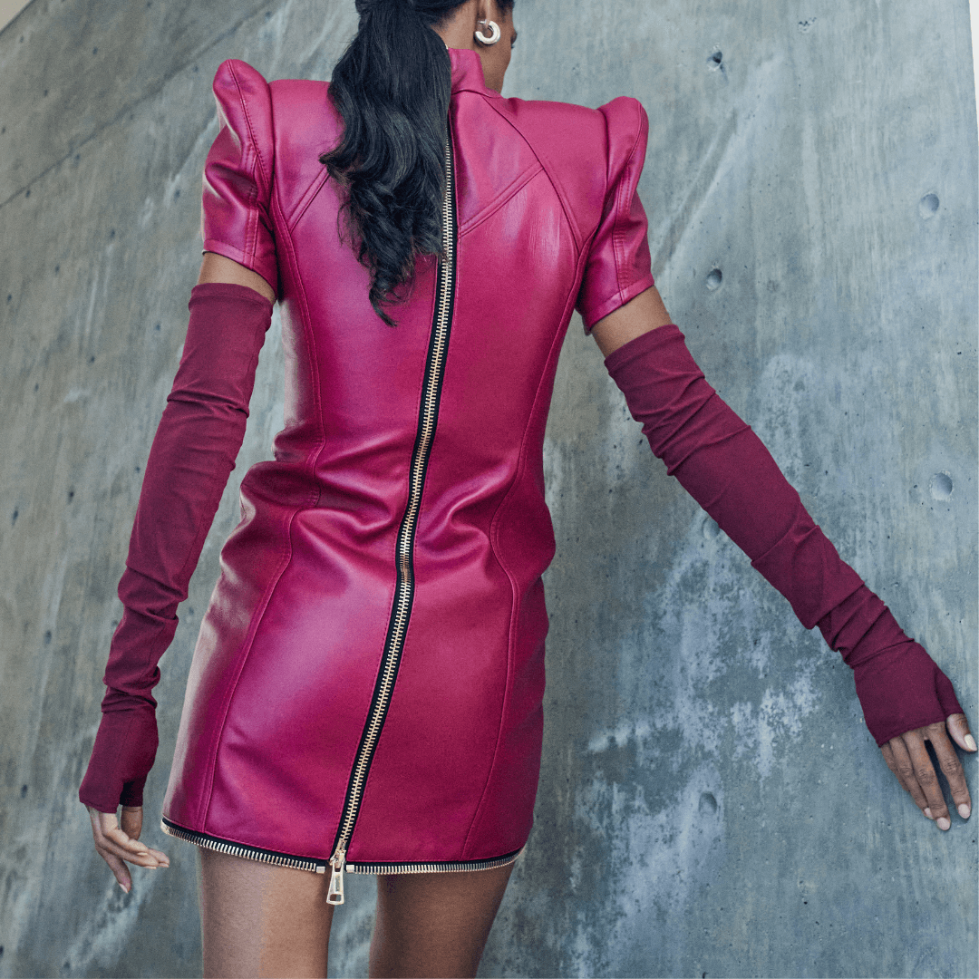 Back view of a woman in a fitted magenta leather mini dress with a full-length zipper and matching fingerless Leigh opera length gloves, standing against a concrete wall.