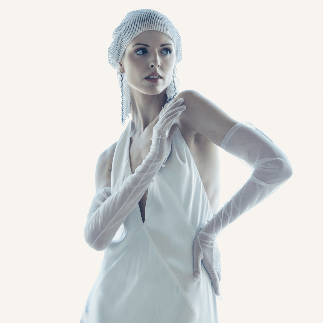 Elegant woman in a white halter gown wearing sheer opera gloves and a crystal mesh headpiece, posing gracefully against a soft white background.