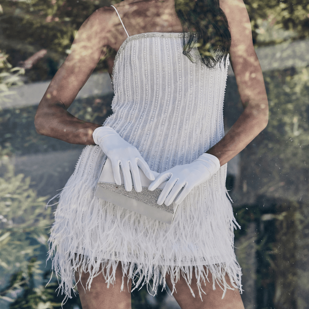 Close-up of a woman wearing a beaded and feathered white mini dress with white gloves, holding a crystal-embellished clutch in front of a reflective glass.
