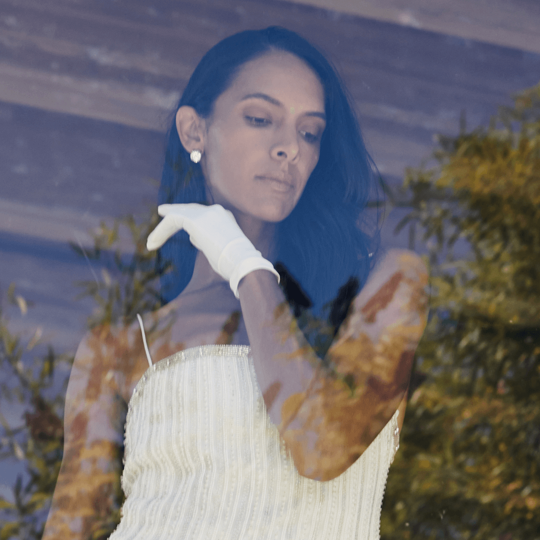 Elegant woman in a beaded white dress and white gloves gazing out a window, with natural reflections of trees and sky softly overlaying the glass.