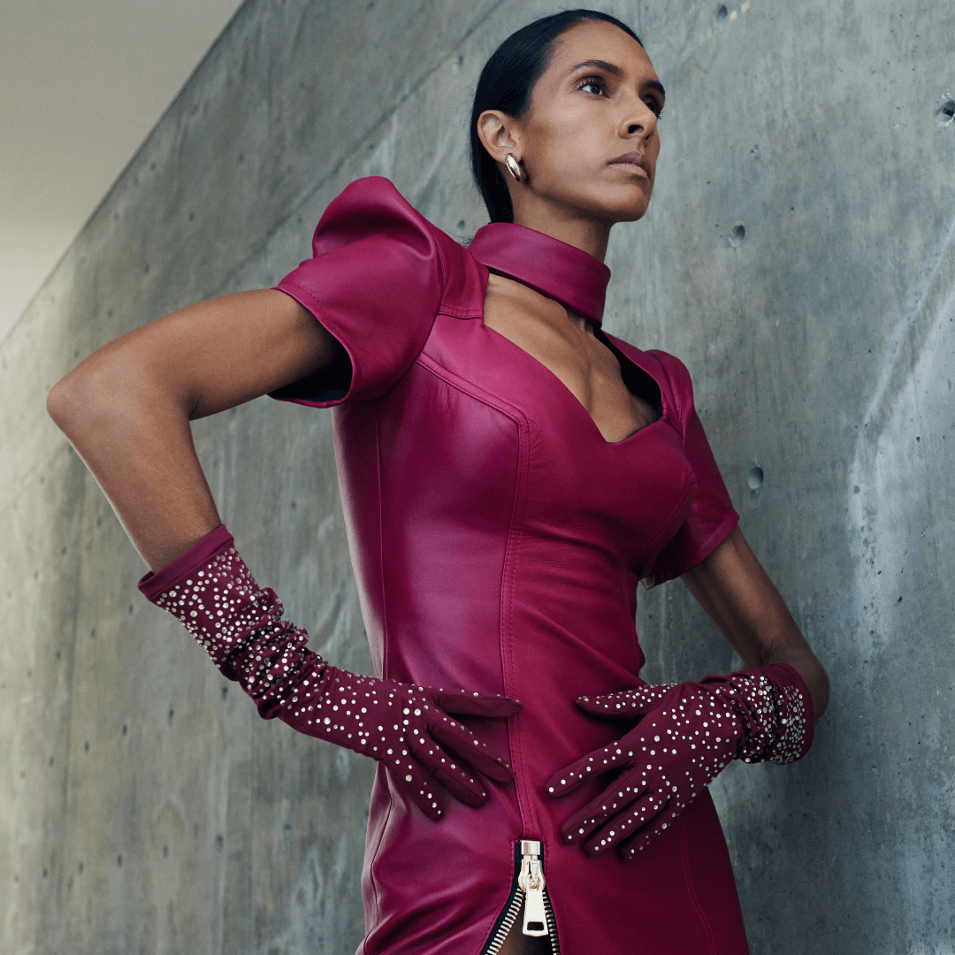 Confident woman in a bold magenta leather dress and matching ruched gloves adorned with crystals, posing against a modern concrete wall backdrop.