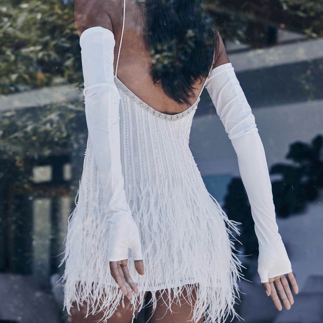 Back view of a woman wearing white satin opera gloves and a white feathered mini dress with pearl embellishments, standing near a reflective glass surface outdoors.