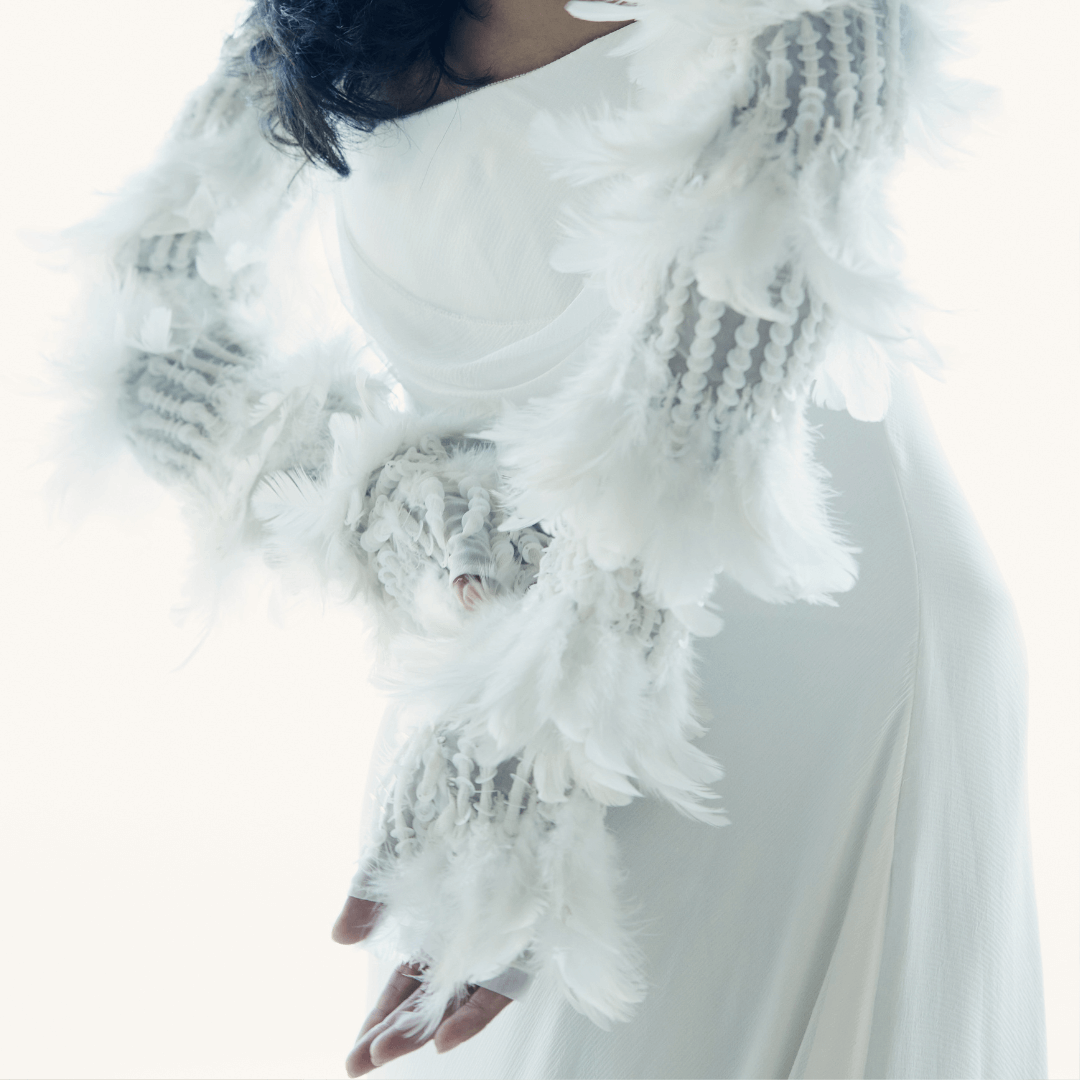 Close-up of a woman in a flowing white gown wearing dramatic white opera gloves adorned with feathers and textured embellishments, captured in soft lighting.