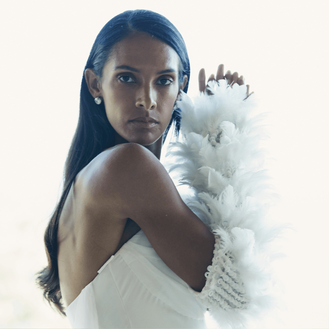 Confident woman in a strapless white gown wearing dramatic feather-adorned opera gloves, posing elegantly in soft natural light.