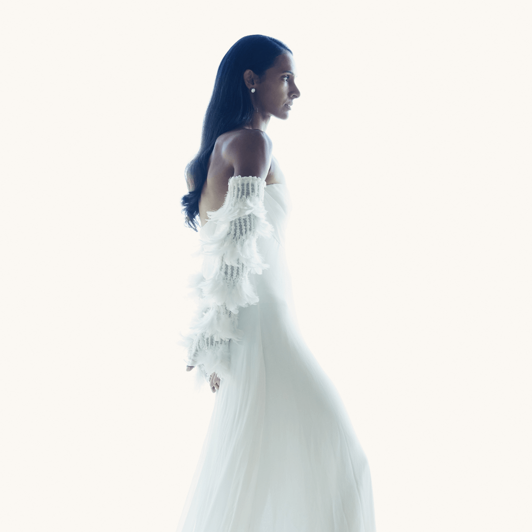 Side profile of a woman in a flowing white gown and dramatic feather-embellished opera gloves, standing gracefully against a bright white backdrop.