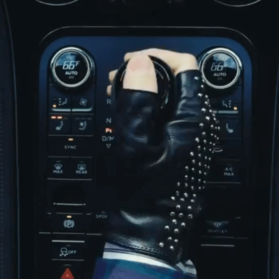 Close-up of a hand wearing black studded fingerless leather gloves gripping a luxury car gear shift, showcasing edgy style and craftsmanship.
