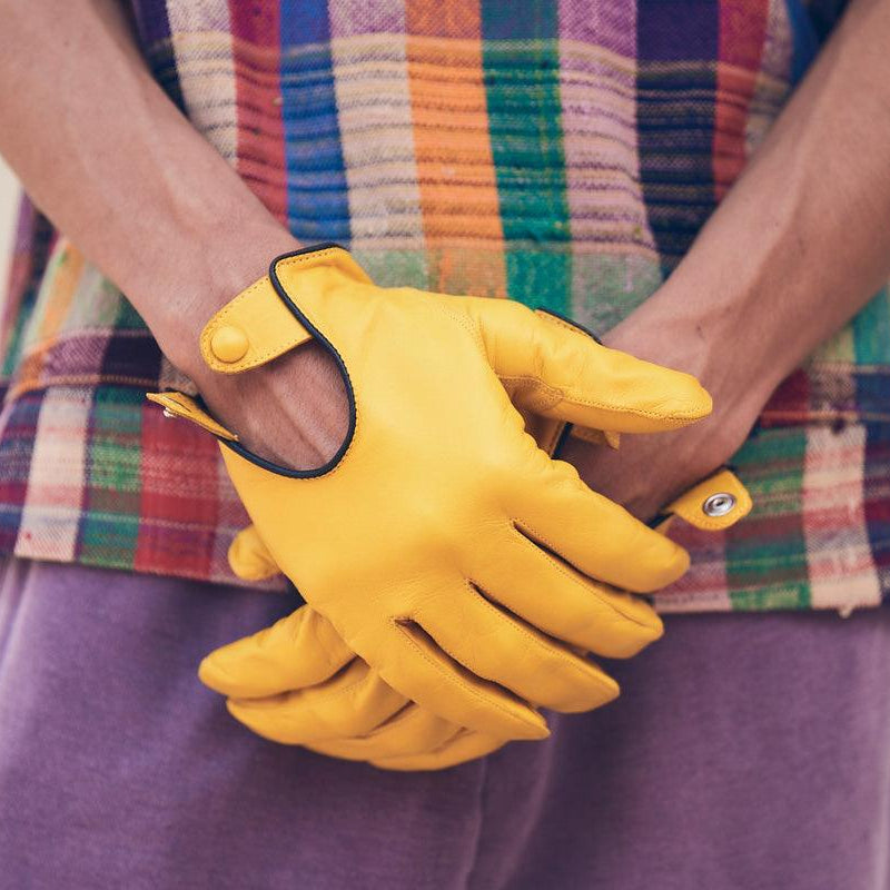 Yellow leather driving gloves with black trim and snap-button closure, worn with a colorful plaid shirt and purple pants.