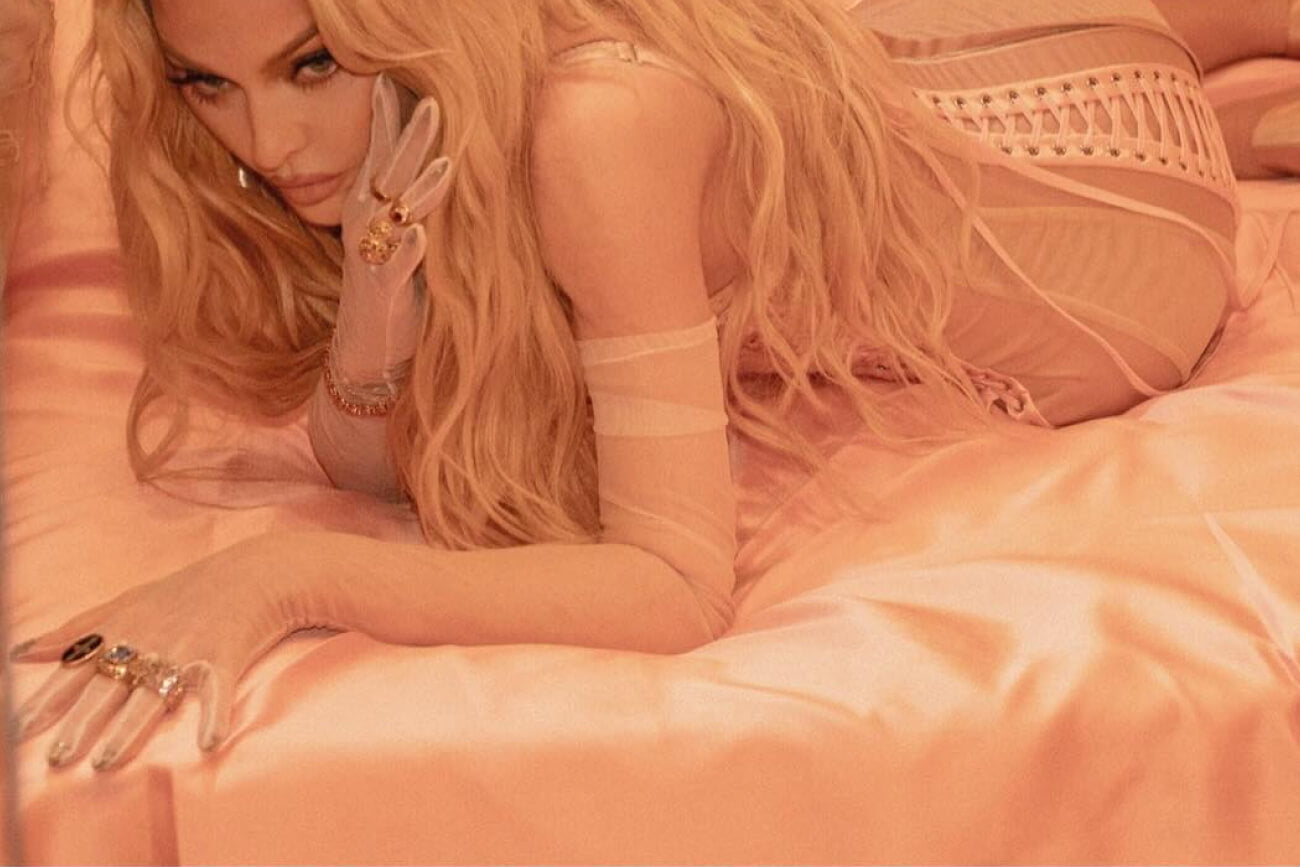 Madonna posing on a satin pink bed, wearing sheer gloves and gold jewelry, with long wavy blonde hair and an intense gaze.
