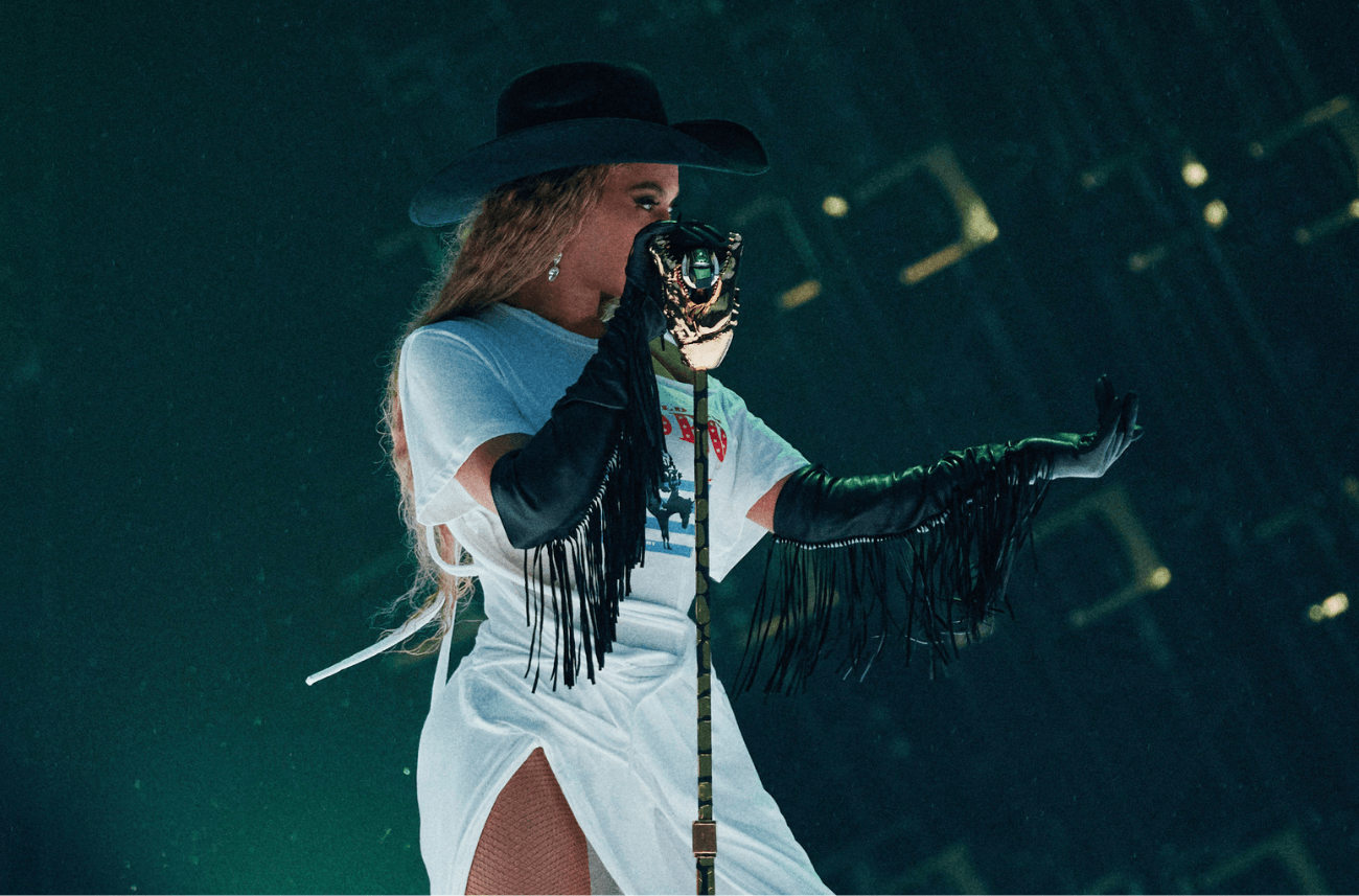 Beyonce wears Seymoure black fringe gloves as she stands on stage wearing a white dress and black cowboy hat