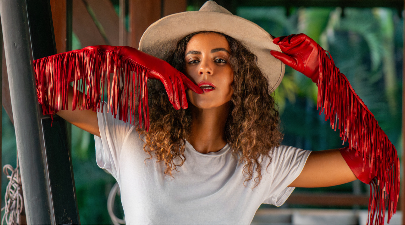 Woman wearing a white t-shirt and beige hat with red fringe gloves, standing against a blurred outdoor background.