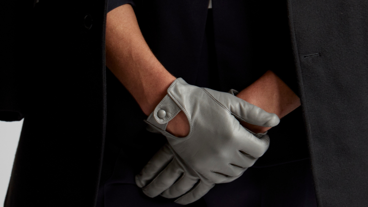 Close-up of a person wearing gray leather driving gloves with a snap-button closure, paired with a dark overcoat for a sleek and sophisticated look.